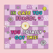 A pastel pink and lavender checkered greeting card with colorful text reading 'IN CASE YOU FORGOT, YOU TOTALLY GOT THIS!' and a white elastic bracelet with a daisy charm laid across the center. The card is photographed against a soft pink background, with 'LETTERBOX LOVE' printed at the bottom.