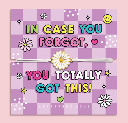 A pastel pink and lavender checkered greeting card with colorful text reading 'IN CASE YOU FORGOT, YOU TOTALLY GOT THIS!' and a white elastic bracelet with a daisy charm laid across the center. The card is photographed against a soft pink background, with 'LETTERBOX LOVE' printed at the bottom.
