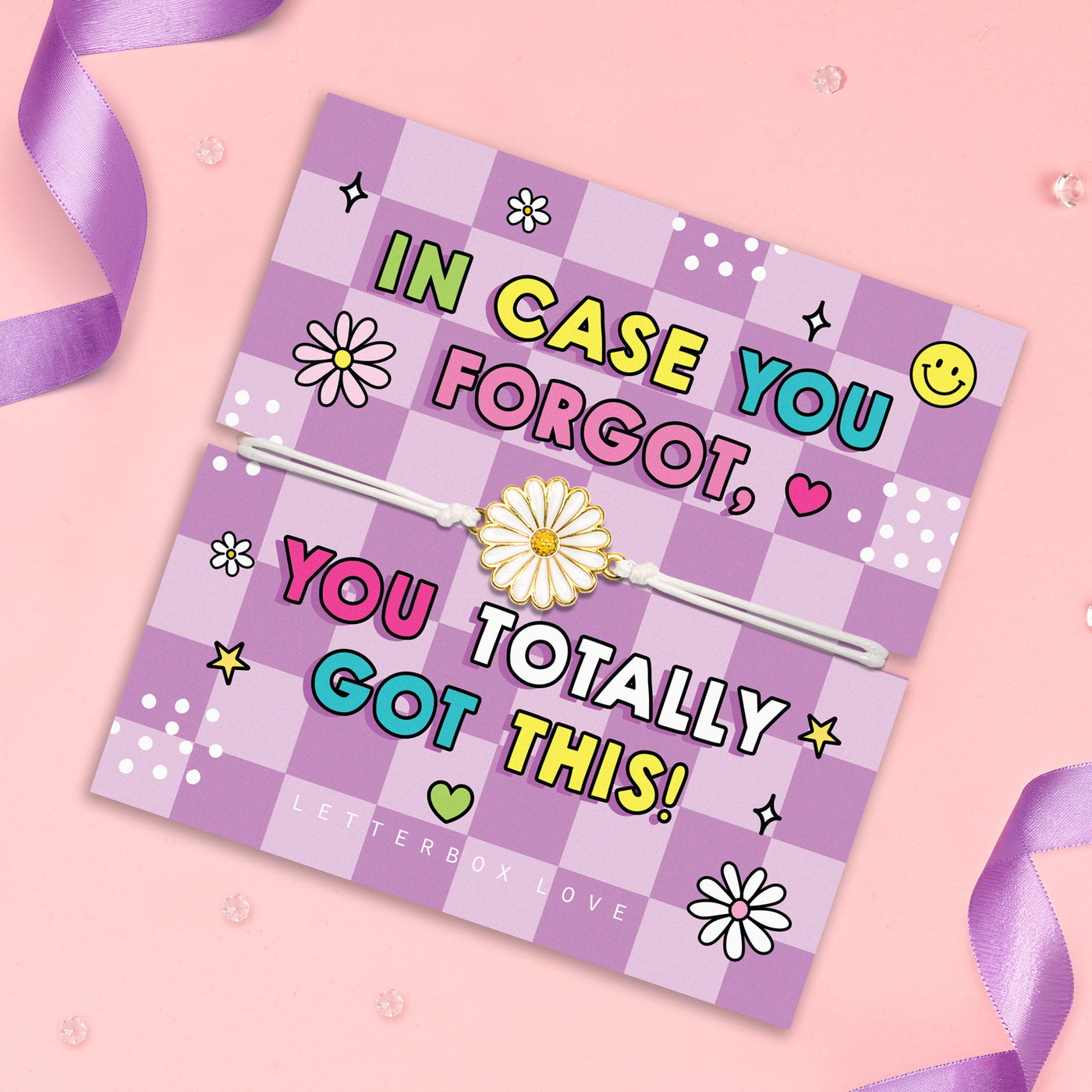 A square lavender and pink checkered letterbox card with a white elastic band across the center, decorated with colorful text reading 'IN CASE YOU FORGOT, YOU TOTALLY GOT THIS!' and small daisy illustrations. The card is photographed on a light pink background with purple ribbon curls and clear decorative beads.