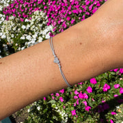 A silver-toned cord bracelet with a small crystal-embellished heart charm, shown worn on a person's wrist against a background of pink and white flowers.