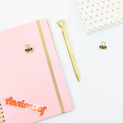 Pink notebook with gold accents, a gold pen, and a small basket on a white background. The journal has a retro 'anxious af' sticker
