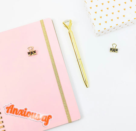 Pink notebook with gold accents, a gold pen, and a small basket on a white background. The journal has a retro 'anxious af' sticker