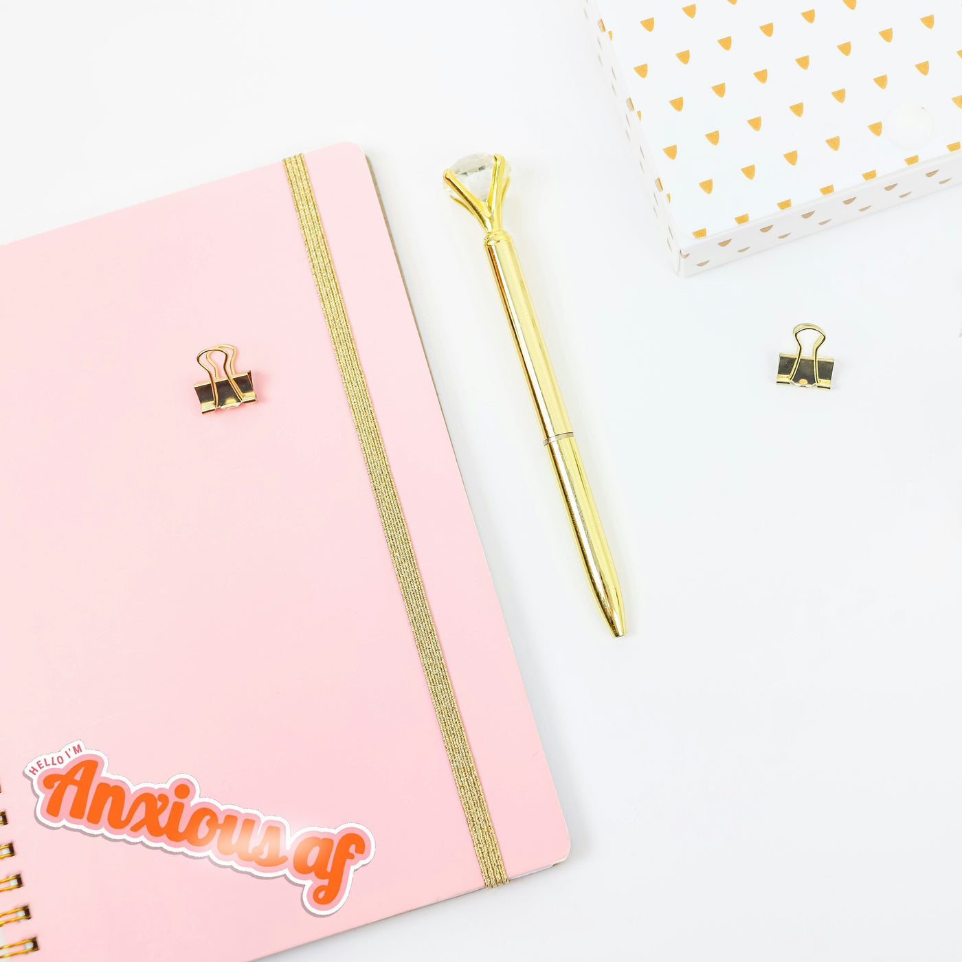 Pink notebook with gold accents, a gold pen, and a small basket on a white background. The journal has a retro 'anxious af' sticker