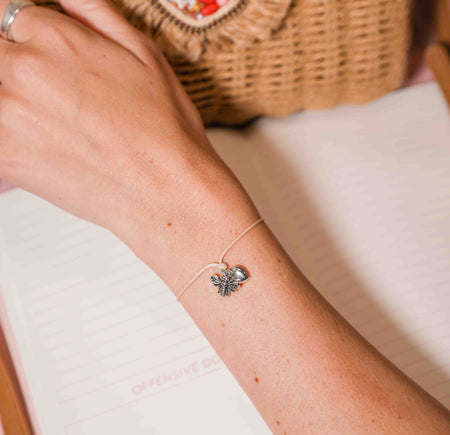 Close-up of a wrist wearing a bracelet with a bee and heart charm, against a neutral background.