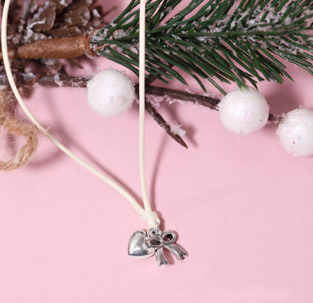A silver heart and bow pendant bracelet on a cream cord, styled with pine branches, white pom-poms, and a cinnamon stick on a solid pink background. A beautiful Christmas gift for her.