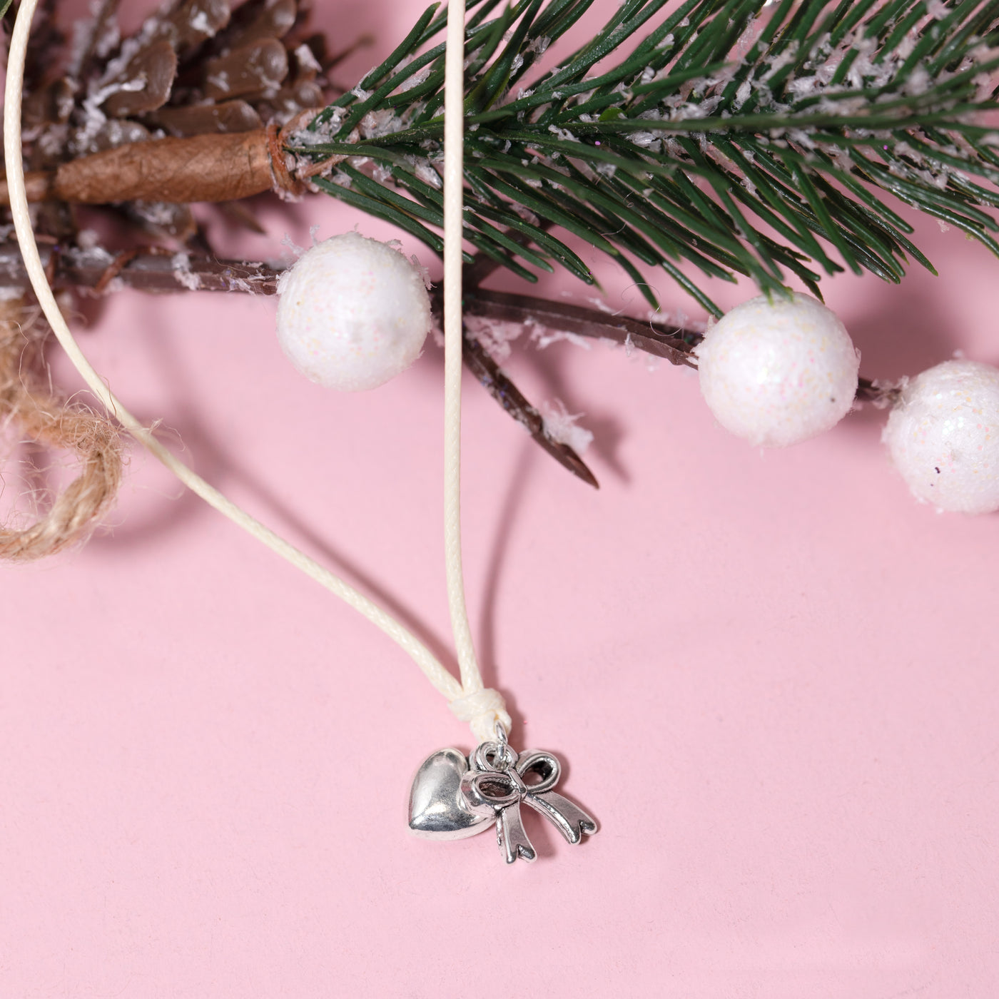 A silver heart and bow pendant bracelet on a cream cord, styled with pine branches, white pom-poms, and a cinnamon stick on a solid pink background. A beautiful Christmas gift for her.