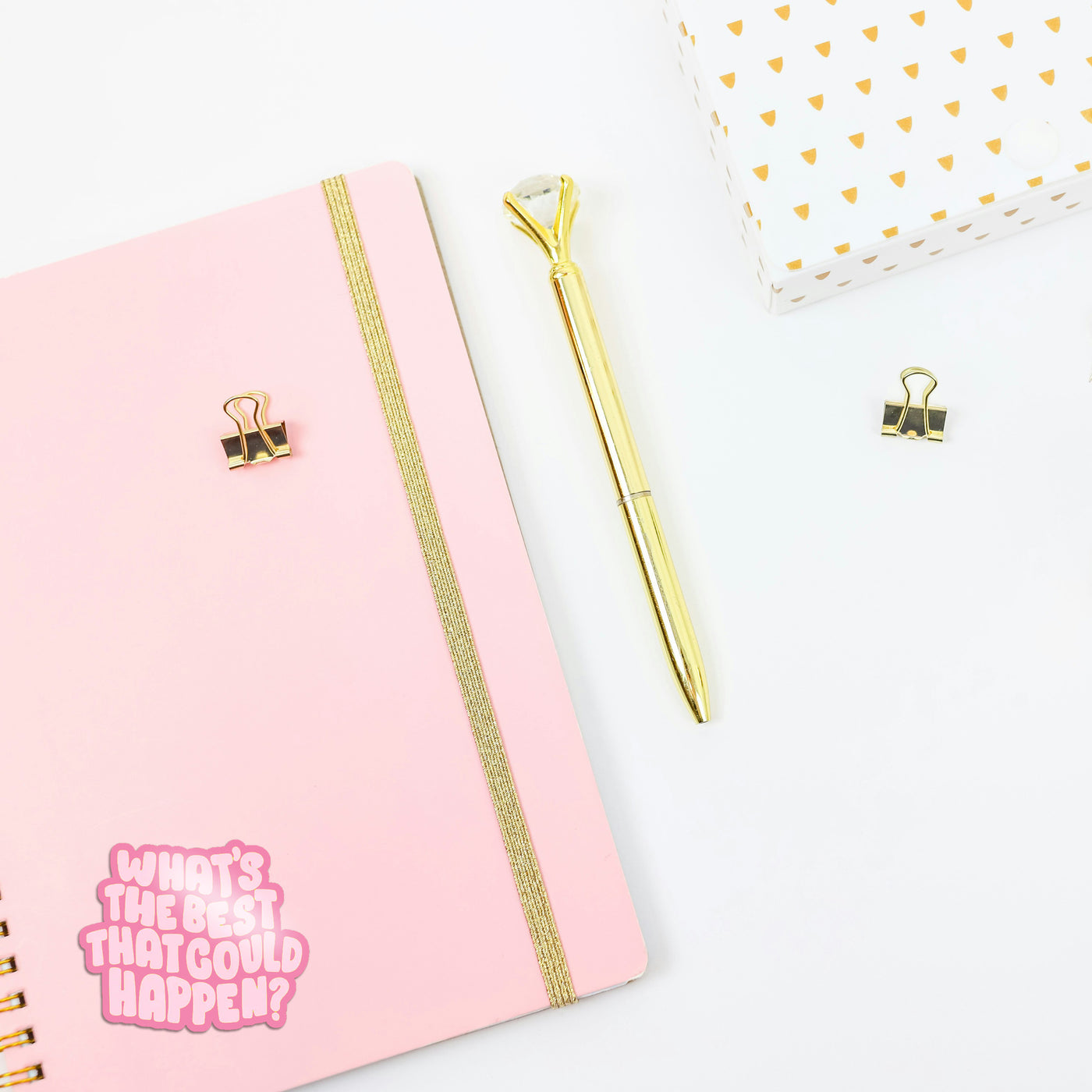Pink notebook with gold accents and a gold pen on a white background. With an aesthetic girly sticker that reads 'whats the best that could happen?'