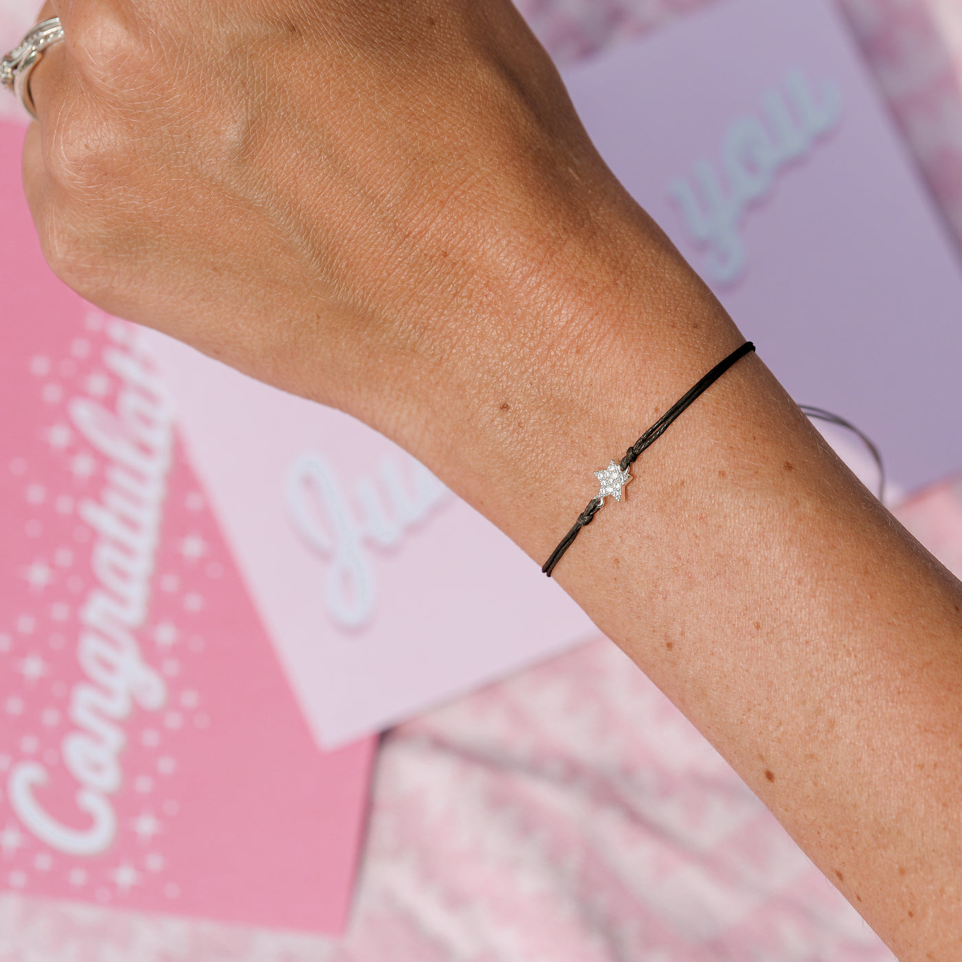 A close-up of a person's wrist wearing a delicate black cord bracelet with a small silver-toned floral charm. Pink and white congratulatory cards are softly blurred in the background.