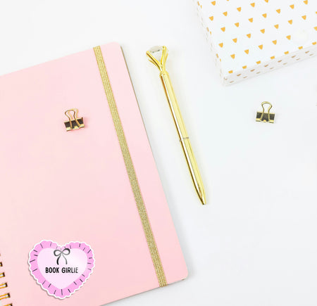 Pink notebook with gold accents, gold pen, and decorative paper clips on a white background. The journal has a 'book girlie' sticker on, perfect as a cute book or kindle sticker.