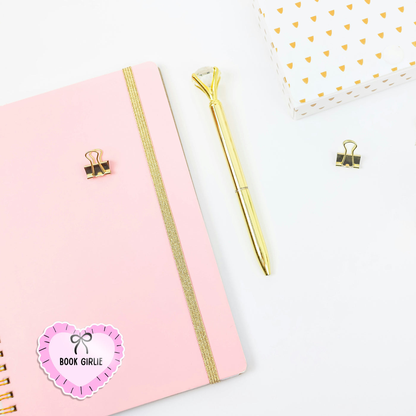 Pink notebook with gold accents, gold pen, and decorative paper clips on a white background. The journal has a 'book girlie' sticker on, perfect as a cute book or kindle sticker.