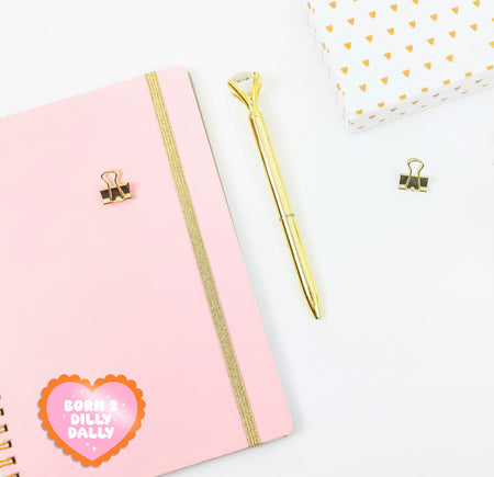Pink notebook with gold accents, a gold pen, and decorative heart-shaped sticker on a white background. On the journal is a pink and orange 'dilly dally' sticker. A cute girly sticker for journals or water bottles