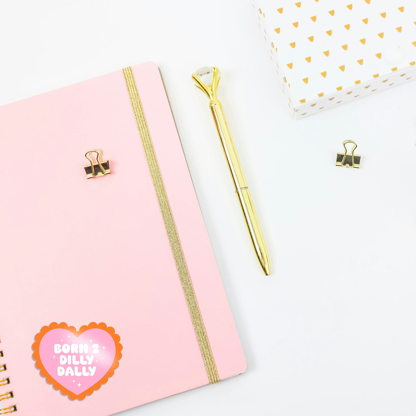 Pink notebook with gold accents, a gold pen, and decorative heart-shaped sticker on a white background. On the journal is a pink and orange 'dilly dally' sticker. A cute girly sticker for journals or water bottles