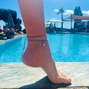 Close-up lifestyle image of a colourful waterproof anklet duo being worn beside a swimming pool on holiday. The adjustable anklets feature pink, turquoise and blue woven cords with a dangling starfish charm, photographed in bright sunshine against a tropical resort backdrop. Perfect summer beach jewellery and holiday accessory.