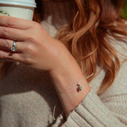 A close-up lifestyle shot of a woman wearing a delicate silver bracelet with a small acorn charm, holding a white coffee cup. She is wearing a beige knit sweater, and her hand with a silver ring is visible in warm sunlight.