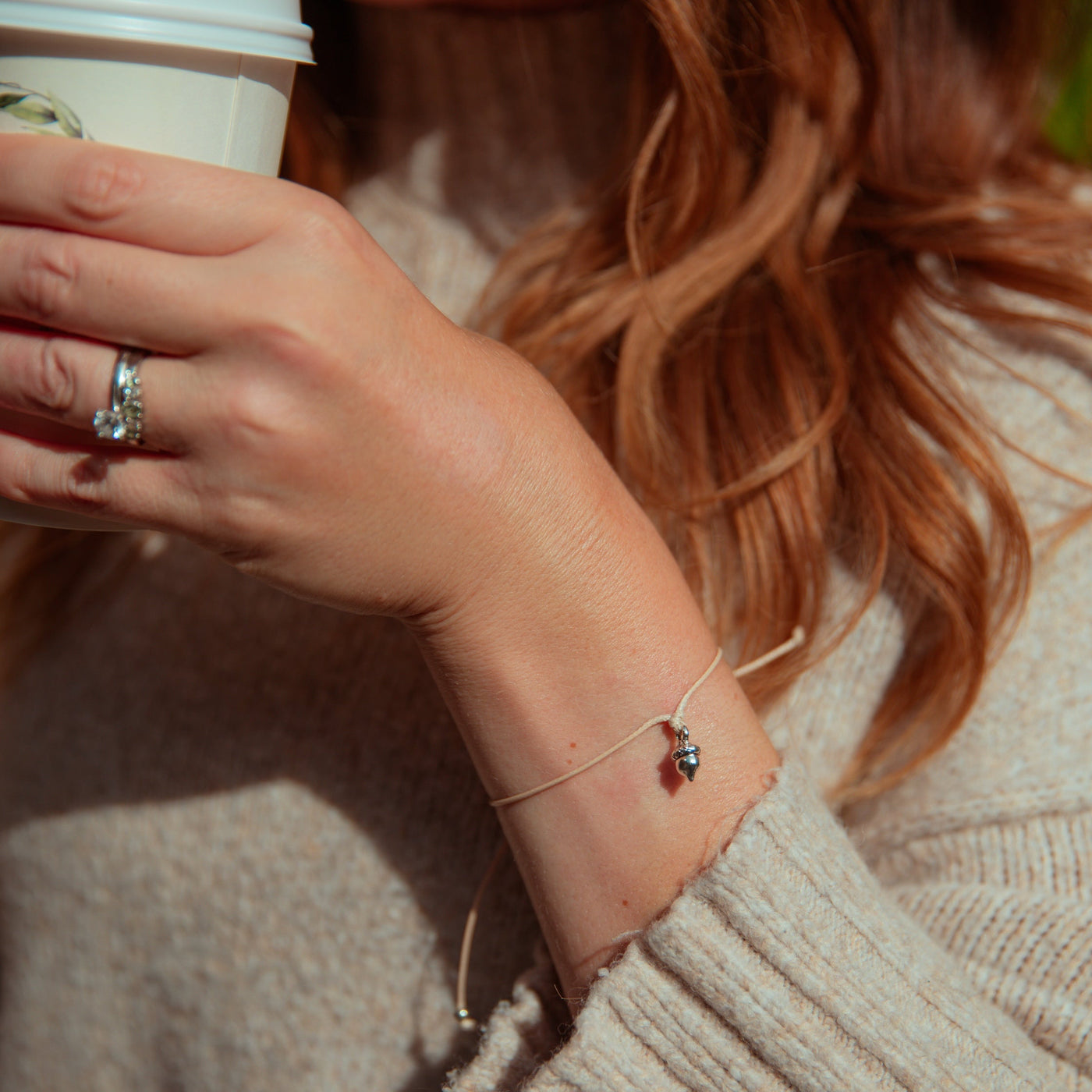 A close-up lifestyle shot of a woman wearing a delicate silver bracelet with a small acorn charm, holding a white coffee cup. She is wearing a beige knit sweater, and her hand with a silver ring is visible in warm sunlight.