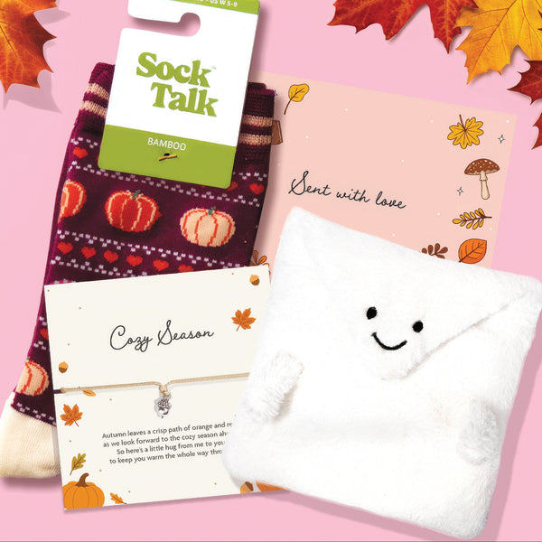 A cozy fall-themed gift set on a pink background with autumn leaves. The set includes a pair of Sock Talk bamboo socks in a dark burgundy knit with orange and white pumpkins, a white plush envelope-shaped pocket with a smiley face, a pink card reading 'Sent with love' decorated with Autumn illustrations and a 'cosy season' bracelet.