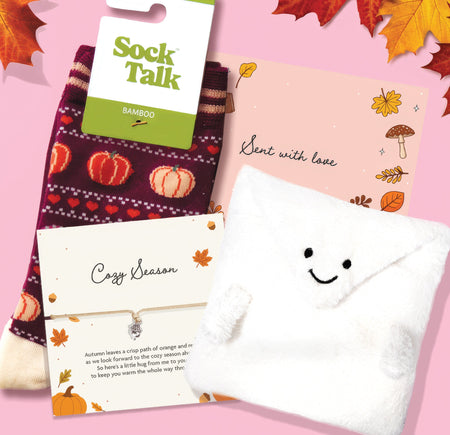 A cozy fall-themed gift set on a pink background with autumn leaves. The set includes a pair of Sock Talk bamboo socks in a dark burgundy knit with orange and white pumpkins, a white plush envelope-shaped pocket with a smiley face, a pink card reading 'Sent with love' decorated with Autumn illustrations and a 'cosy season' bracelet.