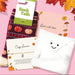 A cozy fall-themed gift set on a pink background with autumn leaves. The set includes a pair of Sock Talk bamboo socks in a dark burgundy knit with orange and white pumpkins, a white plush envelope-shaped pocket with a smiley face, a pink card reading 'Sent with love' decorated with Autumn illustrations and a 'cosy season' bracelet.