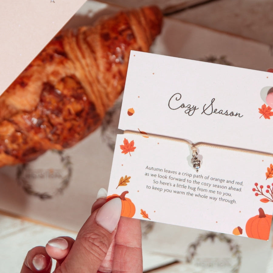 A hand holds a small printed card with autumn-themed illustrations and the words 'Cozy Season' over a coffee cup, croissant, and another matching card on a light tabletop.