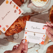 Two hands hold printed autumn-themed cards over a styled tabletop scene with a croissant, whipped cream, and fallen leaves. The cards feature fall illustrations and cursive text, including 'Best with love' and 'Cozy Season.