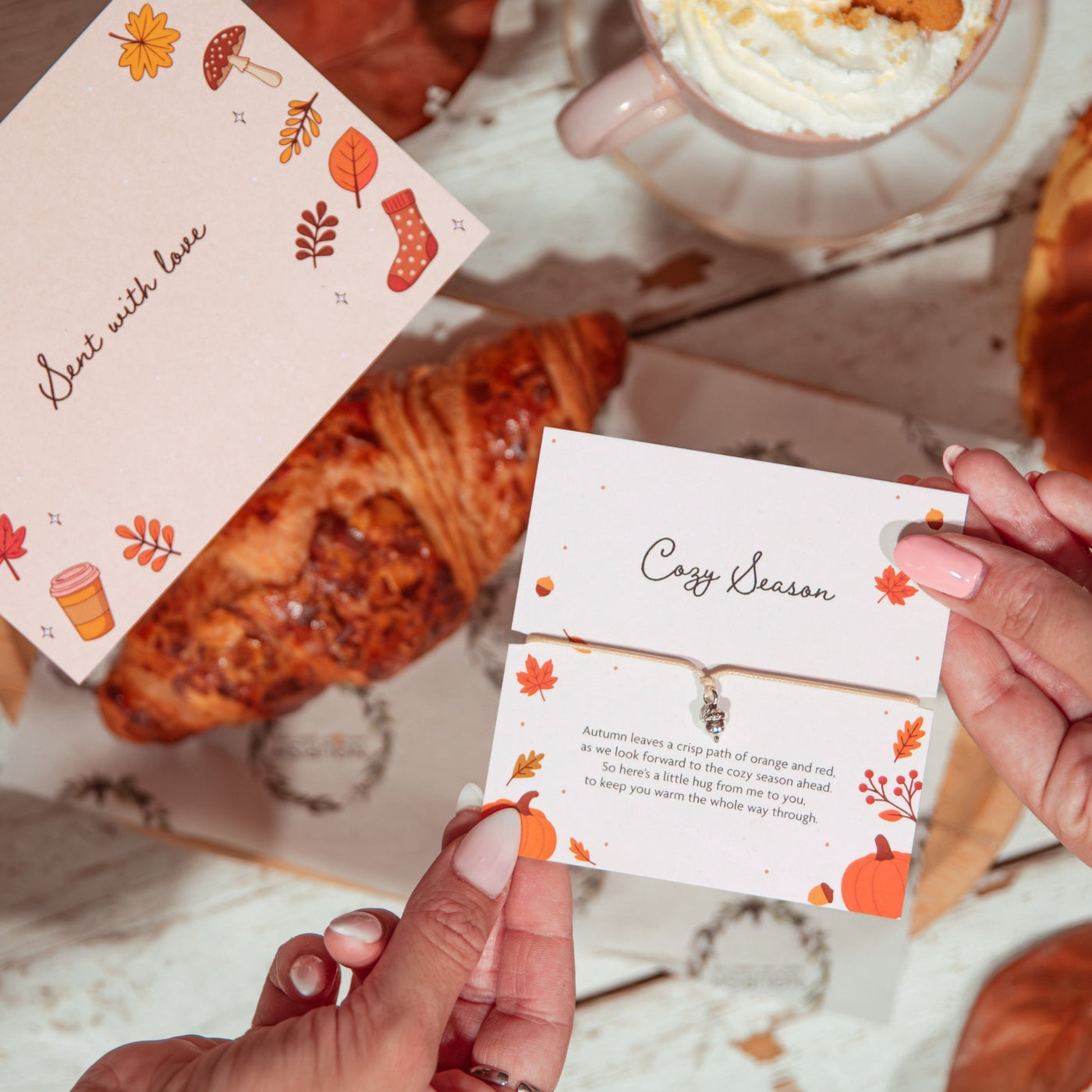 Two hands hold printed autumn-themed cards over a styled tabletop scene with a croissant, whipped cream, and fallen leaves. The cards feature fall illustrations and cursive text, including 'Best with love' and 'Cozy Season.