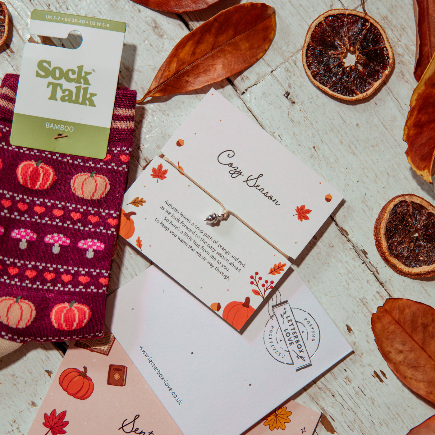 A styled flat-lay of a Sock Talk bamboo socks and a cozy season bracelet on a whitewashed wood background with dried orange slices and autumn leaves. The socks feature a burgundy base with pink, orange, and white knit patterns of pumpkins, hearts, and mushrooms.