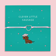 A "Clever Little Sausage" bracelet featuring a silver-plated heart charm on a soft pink adjustable cord. The bracelet is displayed on a teal backing card with an illustration of a dachshund wearing a graduation cap. It's a fun and thoughtful gift for dog lovers and special achievements.