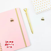 Pink notebook with gold accents and a gold pen on a white background, with a retro style 'in my delulu era' sticker