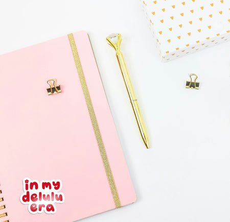 Pink notebook with gold accents and a gold pen on a white background, with a retro style 'in my delulu era' sticker