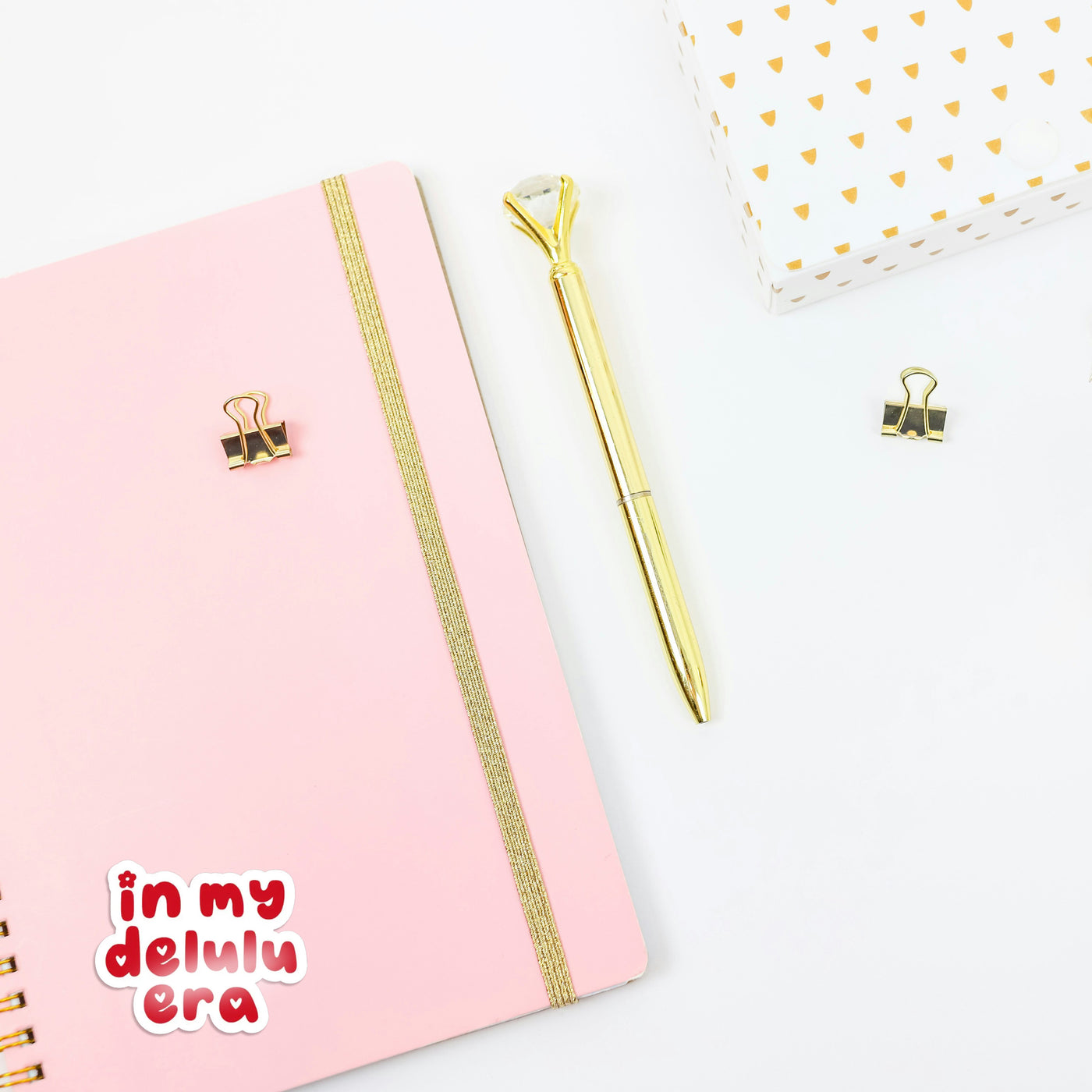 Pink notebook with gold accents and a gold pen on a white background, with a retro style 'in my delulu era' sticker