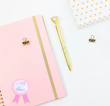 Pink notebook with gold accents and a gold pen on a white background with a 'didnt cry today sticker'