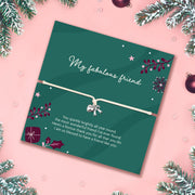 A square green christmas bracelet gift for a friend with a white heart-shaped charm on a bracelet, placed on a pink background with pine branches and ornaments around the edges. The card reads 'My fabulous friend' and includes a festive message and small gift illustration, perfect for a friend at Xmas.
