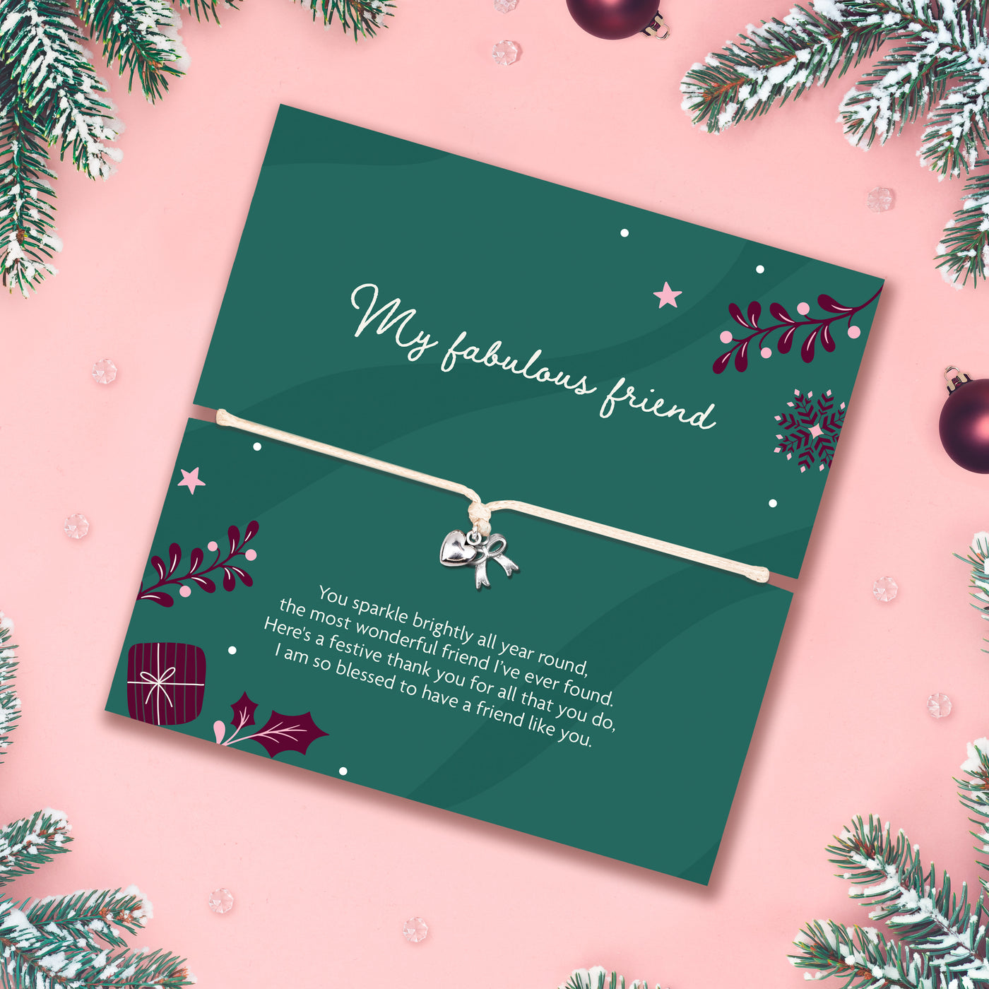 A square green christmas bracelet gift for a friend with a white heart-shaped charm on a bracelet, placed on a pink background with pine branches and ornaments around the edges. The card reads 'My fabulous friend' and includes a festive message and small gift illustration, perfect for a friend at Xmas.
