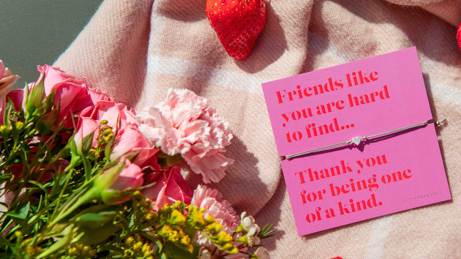 A summer scene with pink blanket, flowers and strawberries. Central is a Letterbox Love friendship bracelet with elegant friendship bracelet. A meaningful gift for a best friend.