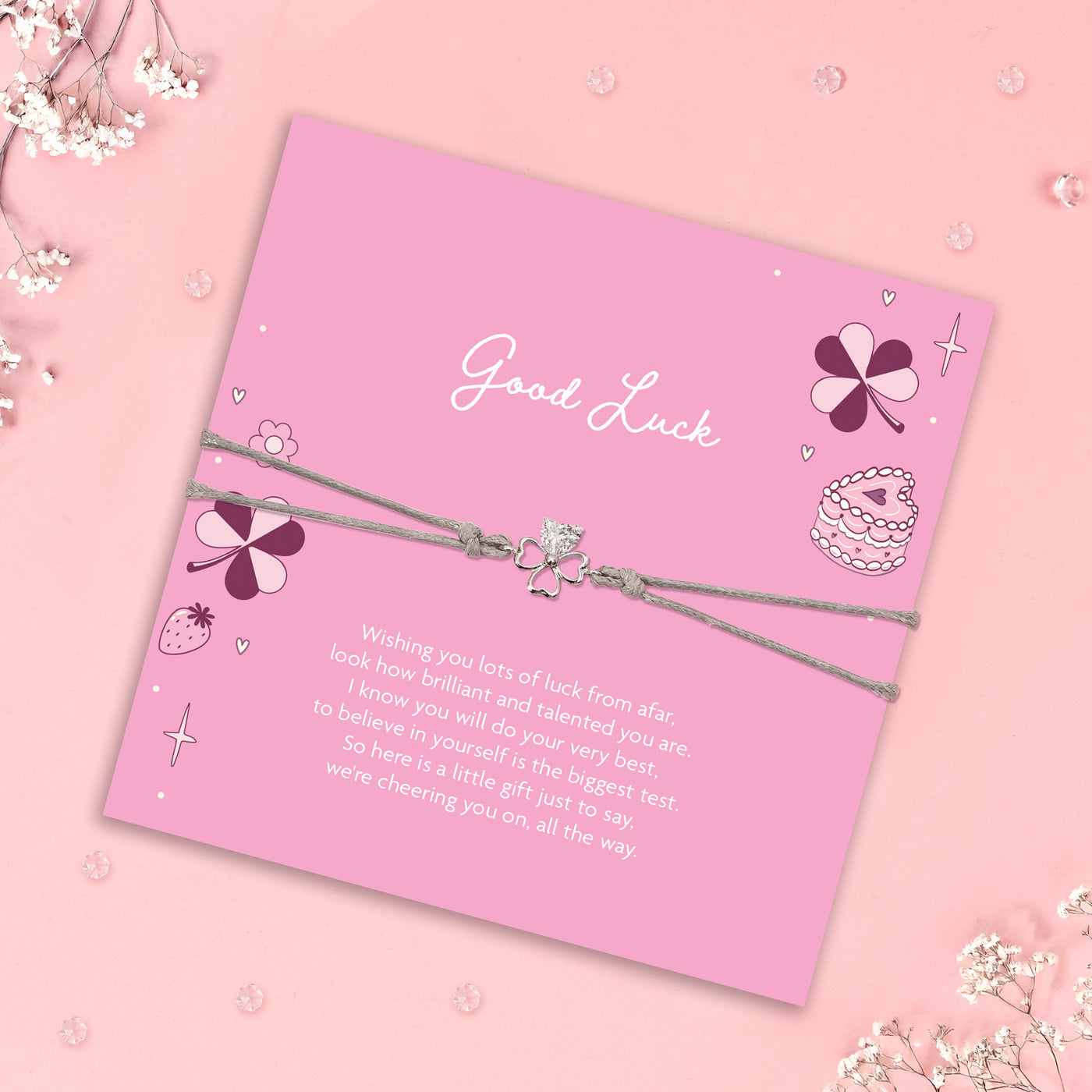 Bracelet with clover charm on a pink card with 'Good Luck' message, surrounded by decorative elements on a pink background.