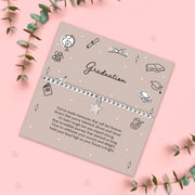Graduation-themed bracelet on a decorative card with green leaves on a pink background, with a silver beaded star bracelet
