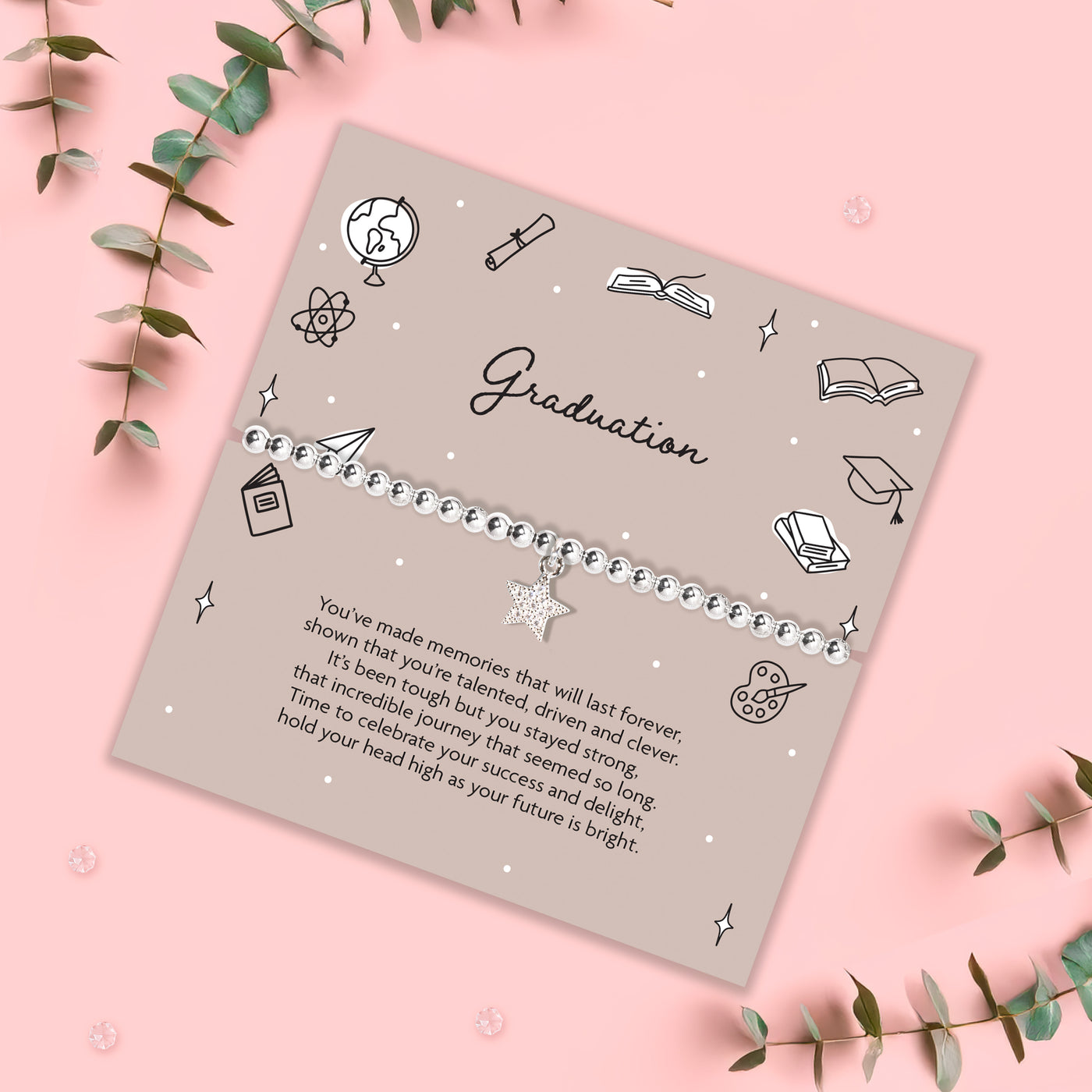 Graduation-themed bracelet on a decorative card with green leaves on a pink background, with a silver beaded star bracelet