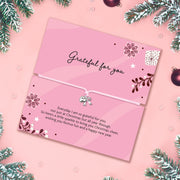 A pink Christmas gift with a white adjustable bracelet and small bow charm, photographed flat on a pink background with pine branches and ornaments around the edges. The card reads 'Grateful for you' and includes a short festive message.