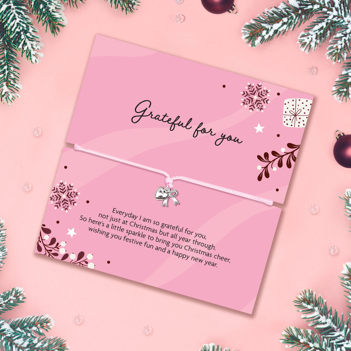 A pink Christmas gift with a white adjustable bracelet and small bow charm, photographed flat on a pink background with pine branches and ornaments around the edges. The card reads 'Grateful for you' and includes a short festive message.