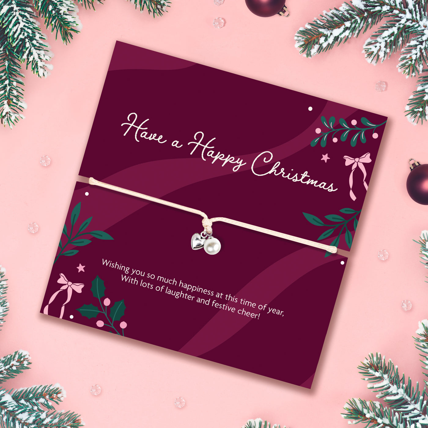 A Christmas-themed poem card featuring a delicate cord bracelet with a silver heart and a pearl charm. The card is deep burgundy with white text reading 'Have a Happy Christmas' and festive green and pink illustrations, set against a light pink background with branches surrounding the card.