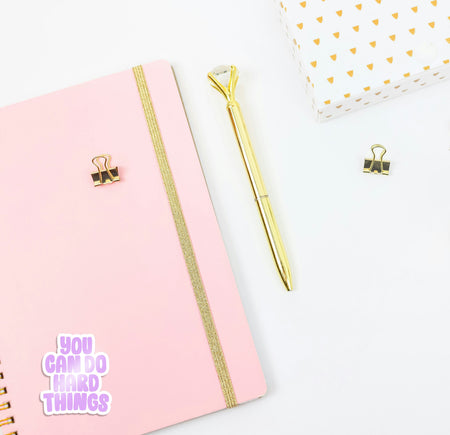 Pink notebook with gold accents and a gold pen on a white background, with a 'you can do hard things' sticker