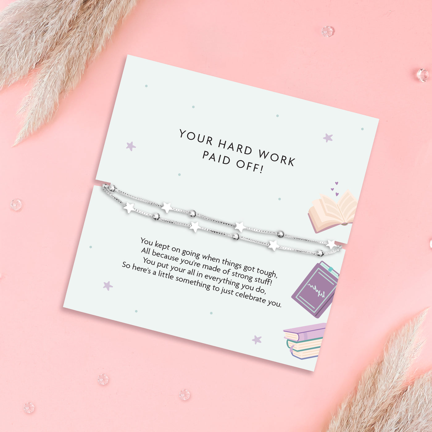 A silver beaded and star charm bracelet displayed on a white printed gift card that reads 'YOUR HARD WORK PAID OFF!' with decorative pastel illustrations, set on a pink background with dried grass accents.