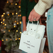 A person in a cream knit sweater holds a white gift bag with the words 'LETTERBOX LOVE' printed on it. A white plush 'Hugsy' gift wrap sits inside the bag, and a blurred Christmas tree with warm lights is in the background.