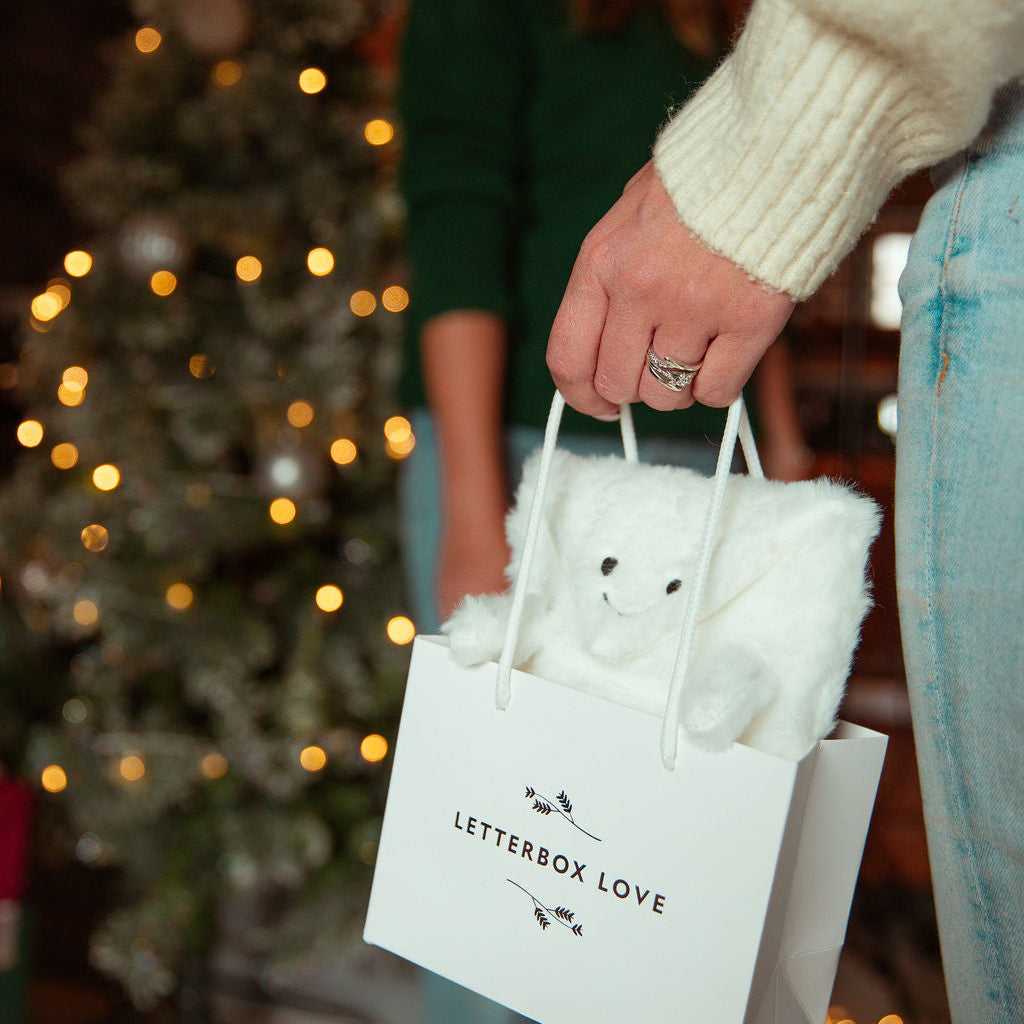 A person in a cream knit sweater holds a white gift bag with the words 'LETTERBOX LOVE' printed on it. A white plush 'Hugsy' gift wrap sits inside the bag, and a blurred Christmas tree with warm lights is in the background.