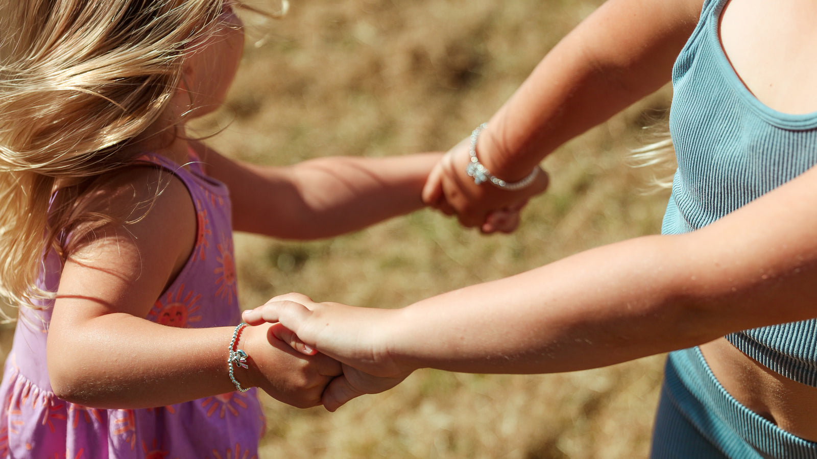 Two little girls holding hands and playing in a field wearing silver-plated beaded bracelets.