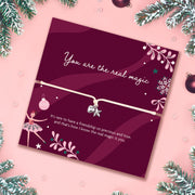 A pink holiday-themed card with a delicate bracelet across the center, surrounded by pine branches and ornaments. The card reads 'You are the real magic' and features ballerina and snowflake illustrations.