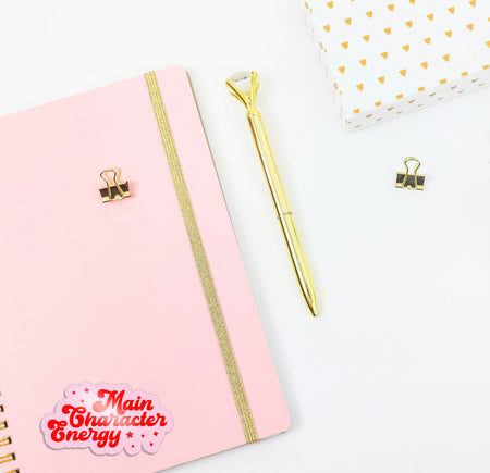 Pink notebook with gold accents and a gold pen on a white background