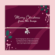 A Christmas gift with a bracelet across the center holding a small silver bow charm. The card is deep burgundy with white script text reading 'Merry Christmas from the bump' and additional holiday illustrations, shown against a light pink background.