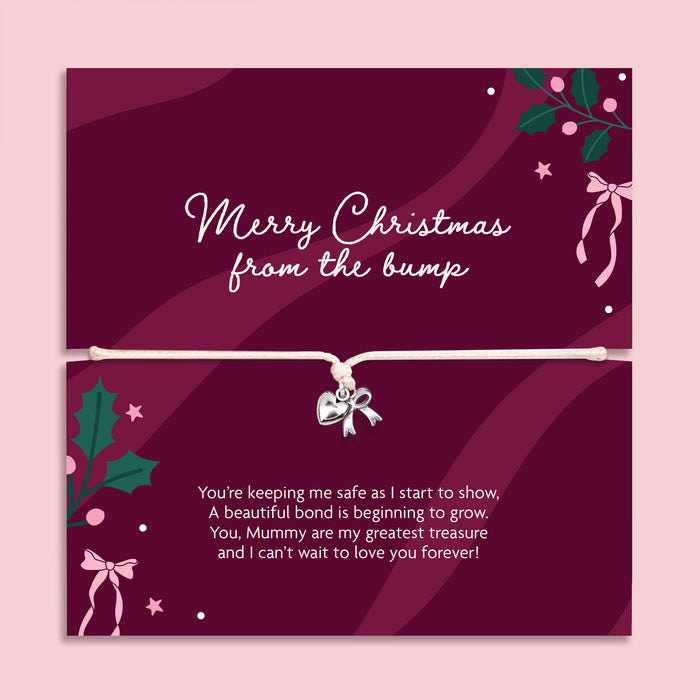 A Christmas gift with a bracelet across the center holding a small silver bow charm. The card is deep burgundy with white script text reading 'Merry Christmas from the bump' and additional holiday illustrations, shown against a light pink background.