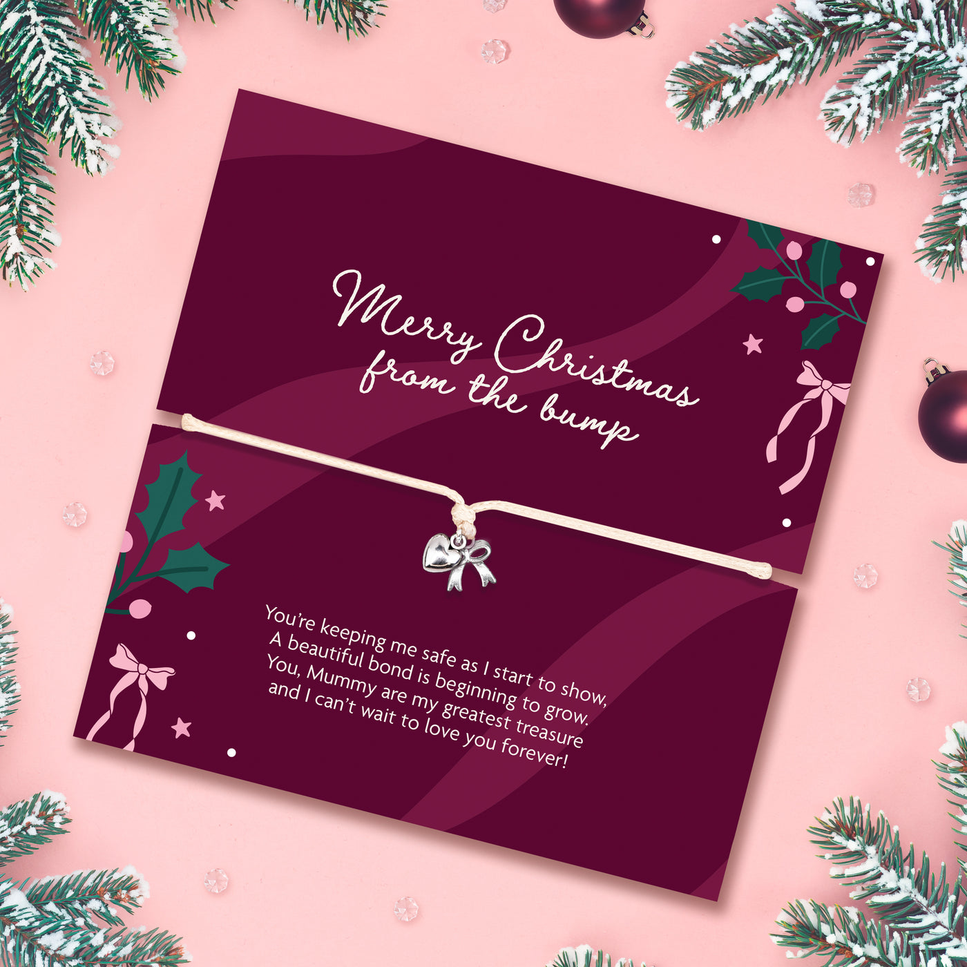A Christmas-themed keepsake card with a white cord bracelet featuring a small silver bow charm, displayed on a pink background with pine branches and ornaments. The card reads 'Merry Christmas from the bump'